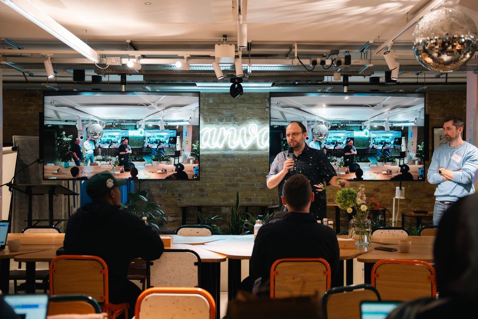 Speaker addresses audience in a modern indoor conference setting, featuring a live presentation on large screens.